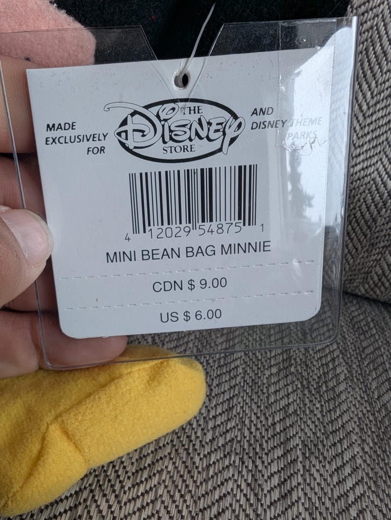 K&ouml;nnte beinhalten: Ein wei&szlig;es Etikett von The Disney Store f&uuml;r einen Minnie Mouse Mini-Beanbag. Das Etikett hat schwarzen Text und einen Barcode. Auf dem Etikett steht "MINI BEAN BAG MINNIE". Das Etikett befindet sich in einer transparenten Kunststoffh&uuml;lle.