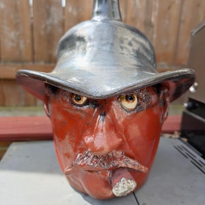 May include: A ceramic sculpture of a face with a wide-brimmed hat, a large nose, and a cigar in its mouth. The face is red, the hat is gray, and the cigar is white.