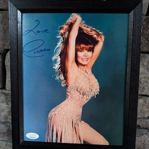 May include: A framed photograph of a woman with long red hair wearing a sequined dress. The woman is smiling and holding her hair up. The photograph is signed "Love,  [illegible]" in blue ink.