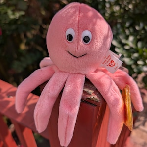 May include: A pink plush octopus toy with large, round eyes and a smiling mouth. The toy has eight tentacles and a tag that reads "Inky" from the Beanie Babies Collection. The octopus is sitting on a red wooden surface.