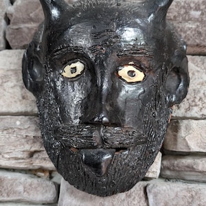 May include: A black ceramic mask with a beard and horns. The mask has two yellow eyes and a textured surface.
