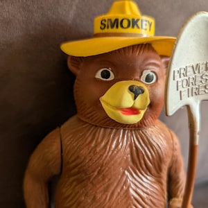 Puede incluir: Figura de Smokey Bear marrón con un sombrero amarillo con la inscripción "SMOKEY". El oso sostiene una pala con las palabras "PREVENT FOREST FIRES". La figura tiene un hocico amarillo y una boca roja.