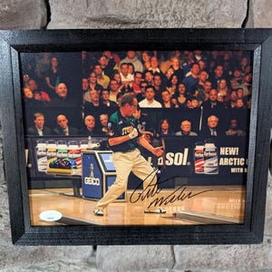 May include: A framed photograph of a man in a green bowling shirt, holding a bowling ball, with a crowd of people in the background. The photo is signed by the man, "Pete Weber".