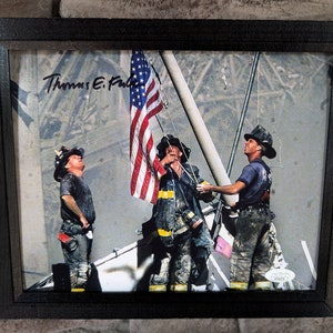 May include: A framed photograph of three firefighters raising an American flag on a flagpole against a backdrop of smoke and debris. The firefighters are wearing helmets and fire-resistant clothing. The photograph is signed "Thomas E.  Fulton".