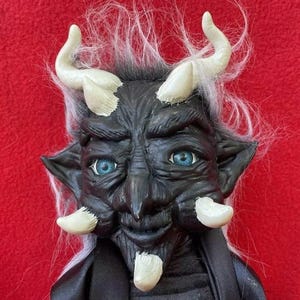 May include: A black and white goblin doll with long white hair and horns. The doll has blue eyes and a mischievous grin.