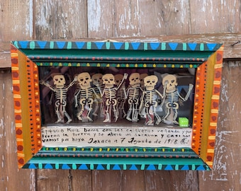 May include: A framed folk art piece featuring a group of skeleton figures. The frame is painted with orange, green, and blue accents. The skeletons are white with black details, and the piece includes handwritten text.