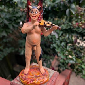May include: A handcrafted figurine of a devil playing a violin. The figure has horns, wings, and a tail, standing on a fiery base. The violin is brown and the figure is painted in flesh tones.