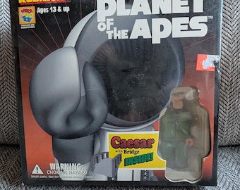 Könnte beinhalten: Eine schwarz-weiße Box mit einer Kubrick Planet der Affen Figur im Inneren. Die Box hat einen Warnhinweis über Erstickungsgefahr. Die Figur ist Caesar, eine Figur aus der Planet der Affen Reihe. Die Box enthält auch eine kleine Brücke.