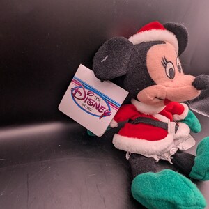 K&ouml;nnte beinhalten: Ein Minnie Mouse Pl&uuml;schtier mit Weihnachtsm&uuml;tze und rot-wei&szlig;em Outfit. Das Pl&uuml;schtier hat schwarze Ohren, ein rosa Gesicht und gr&uuml;ne Schuhe. Ein Disney Store-Etikett ist angebracht.