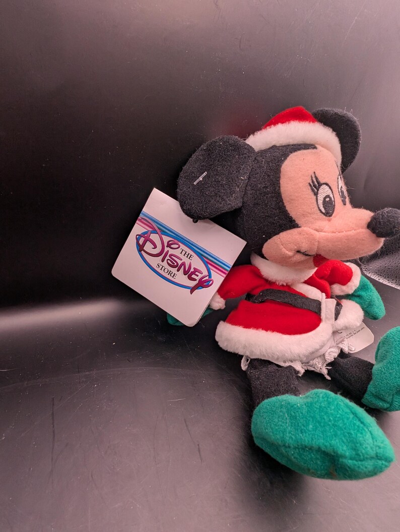 K&ouml;nnte beinhalten: Ein Minnie Mouse Pl&uuml;schtier mit Weihnachtsm&uuml;tze und rot-wei&szlig;em Outfit. Das Pl&uuml;schtier hat schwarze Ohren, ein rosa Gesicht und gr&uuml;ne Schuhe. Ein Disney Store-Etikett ist angebracht.