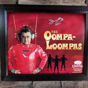 May include: A framed poster featuring a character from the film "Charlie and the Chocolate Factory." The character is wearing a red jumpsuit and a headset. The poster has the text "The Oompa-Loompas" and a signature at the top.