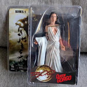 May include: A Queen Gorgo action figure from the movie 300, in a clear plastic package. The figure is wearing a white dress with gold accents. The packaging includes the movie's logo and the text "Queen Gorgo". A Series 1 card is visible.