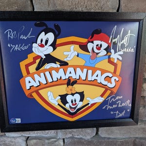 Op de afbeelding: Ingelijste Animaniacs-print met Yakko, Wakko en Dot, met het logo van de show. De print staat tegen een blauwe achtergrond in een zwart frame. De handtekeningen van Rob Paul, Jess Harnell en Tress MacNeille zijn zichtbaar.
