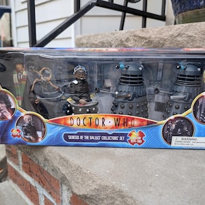 May include: A Doctor Who Genesis of the Daleks collectors set in a rectangular box. The set includes a Fourth Doctor figure, two Dalek figures, a Davros figure, and a baby mutant Dalek accessory. The packaging features blue graphics and text.