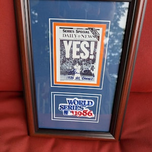 May include: Framed print featuring a newspaper headline that reads "YES!" with the text "SERIES SPECIAL DAILY NEWS." Below is a "WORLD SERIES 1986" logo. The frame is dark brown with a blue matting.