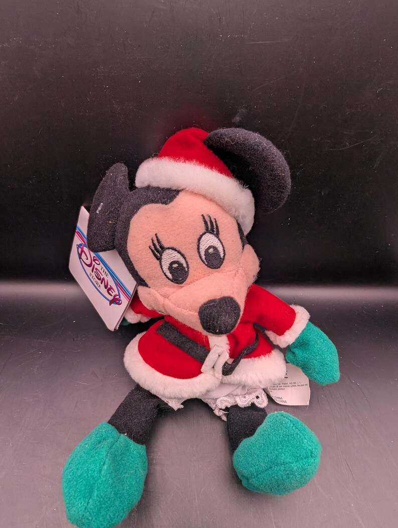 K&ouml;nnte beinhalten: Ein Pl&uuml;sch Minnie Mouse-Puppe in einem Weihnachtsmann-Outfit. Die Puppe hat einen rot-wei&szlig;en Weihnachtsmannhut, einen roten Mantel mit wei&szlig;em Besatz und gr&uuml;ne Handschuhe und Schuhe. Das Disney Store-Etikett ist angebracht.