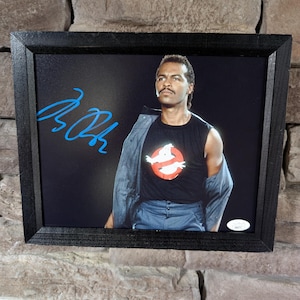 May include: Framed photograph featuring a man wearing a black Ghostbusters t-shirt with the iconic logo. The image is signed in blue ink. The frame is black and the background is a dark color.
