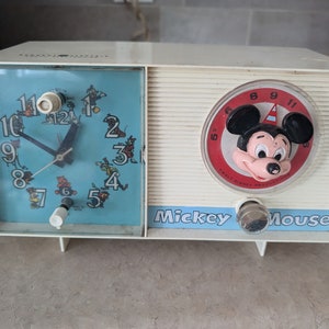 May include: Vintage white Mickey Mouse clock radio with a blue face and Mickey Mouse's face on the front. The clock face has a blue background with black hands and white numbers. The radio has a red and white dial with Mickey Mouse's face on it.