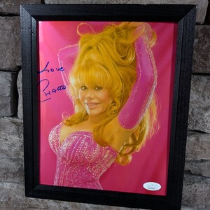 May include: A framed photograph of a woman with blonde hair wearing a pink sequined dress and pink gloves. The woman is smiling and looking at the camera. The photo is signed "Love,  Hara."