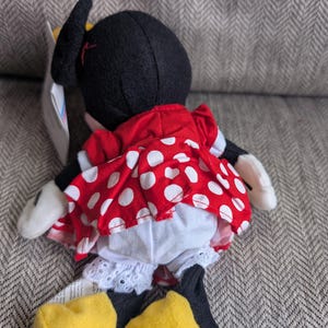 Puede incluir: Mu&ntilde;eca de peluche de Minnie Mouse, vista desde atr&aacute;s, con un vestido rojo con lunares blancos y bragas blancas con ribete de encaje. La mu&ntilde;eca tiene orejas negras, zapatos amarillos y guantes blancos. Una etiqueta est&aacute; adjunta.
