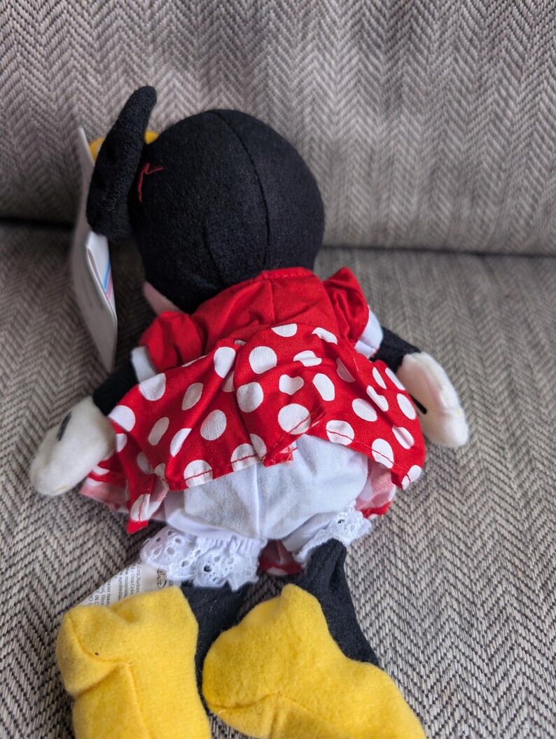 Puede incluir: Mu&ntilde;eca de peluche de Minnie Mouse, vista desde atr&aacute;s, con un vestido rojo con lunares blancos y bragas blancas con ribete de encaje. La mu&ntilde;eca tiene orejas negras, zapatos amarillos y guantes blancos. Una etiqueta est&aacute; adjunta.