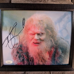 以下が含まれることがあります： 顔の周りに赤い光を放つ、長い白いひげを生やした男性の白黒写真が額縁に入っています。男性は不気味な笑顔でカメラを見つめています。写真は黒いペンでサインされています。