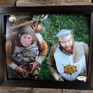 May include: A framed photograph of two men dressed as medieval knights. The man on the left is wearing a brown tunic and a helmet. The man on the right is wearing a chainmail tunic and a crown. The photograph is signed by both men.