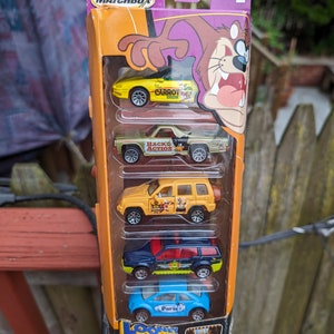 Könnte beinhalten: Ein Matchbox-Autoset mit fünf Die-Cast-Fahrzeugen aus der Looney Tunes-Zeichentrickserie. Das Set enthält einen gelben Wagen mit der Aufschrift "The Carrot Crew", einen roten Pickup mit der Aufschrift "Back in Action", einen gelben Geländewagen mit der Aufschrift "The Wild Bunch", einen blauen Geländewagen mit der Aufschrift "The Road Runners" und einen blauen Volkswagen Käfer mit der Aufschrift "Paris". Das Set ist in einer Pappbox mit einem Bild von Bugs Bunny und einer Zeichnung des Tasmanian Devils verpackt.