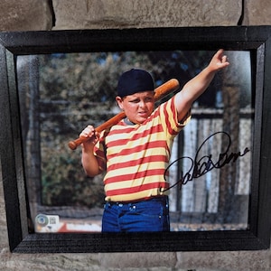 May include: Framed photograph featuring a young boy in a yellow and red striped shirt, blue jeans, and a black cap, holding a baseball bat. The image is signed in black ink. The frame is black.