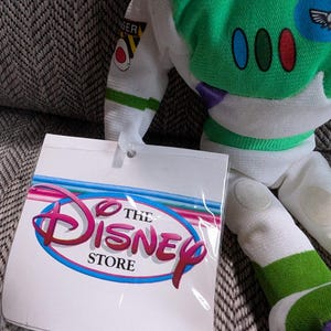 Op de afbeelding: Een Buzz Lightyear pluche pop met een groen en wit ruimtepak. De pop heeft een label van The Disney Store. Het label heeft het Disney-logo in roze en rood met een blauwe omlijning.