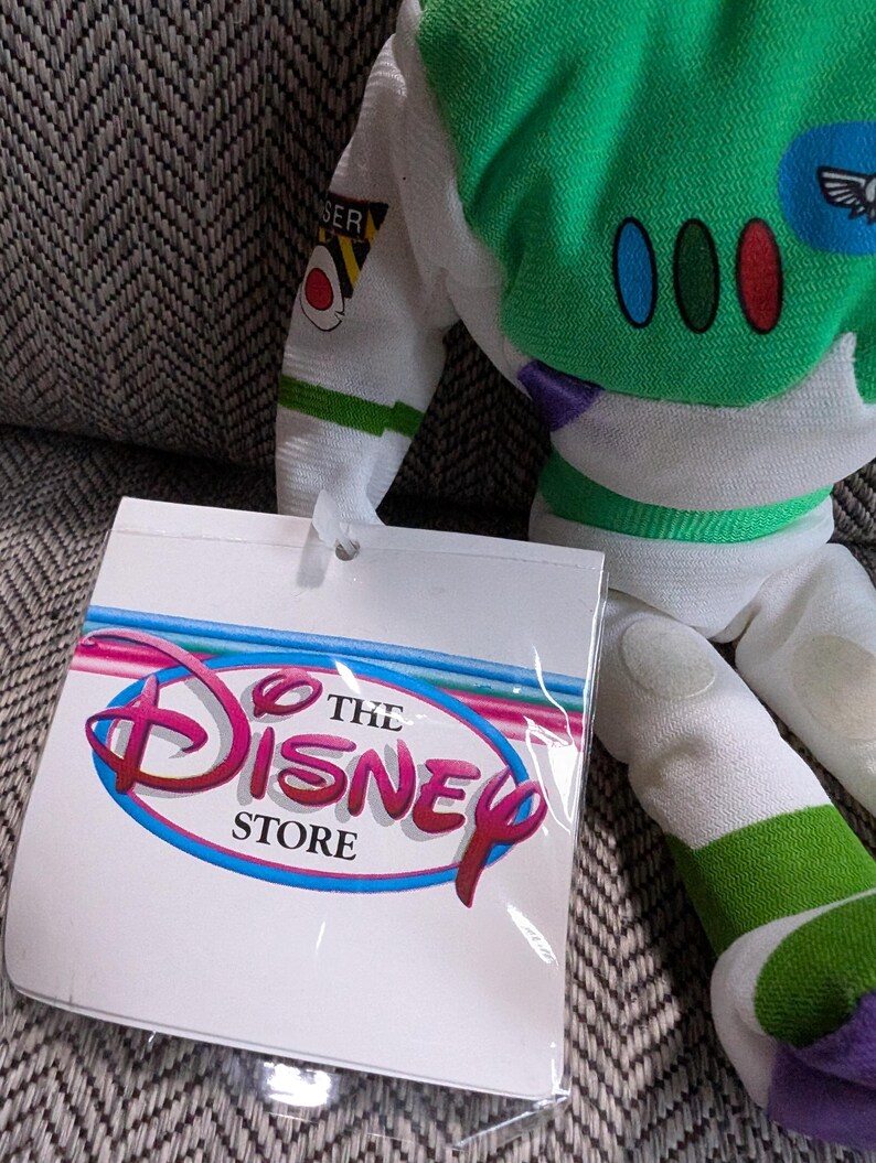 Op de afbeelding: Een Buzz Lightyear pluche pop met een groen en wit ruimtepak. De pop heeft een label van The Disney Store. Het label heeft het Disney-logo in roze en rood met een blauwe omlijning.