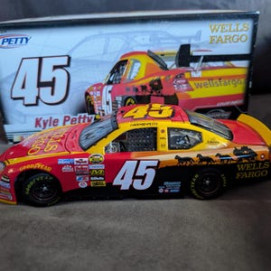 Op de afbeelding: Een rood, geel en zwart Wells Fargo #45 Kyle Petty raceauto model. De auto heeft nummer 45 op de zijkant en het dak. De doos achter de auto toont ook het ontwerp van de auto en het Wells Fargo logo.