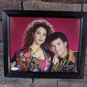 May include: Framed photograph featuring two people, a woman with curly hair and a man, both smiling. The photograph is set against a pink background and is signed in gold ink. The frame is black.