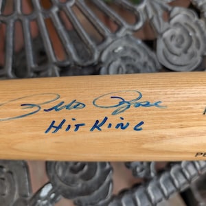 May include: A light-colored wooden baseball bat with blue ink signatures and the words "Hit King". The bat has the word "ADIR" printed on it.