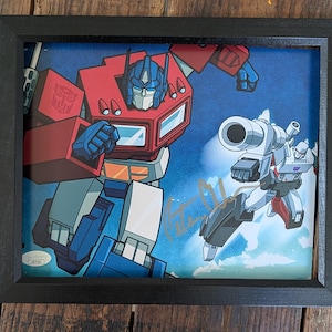 May include: A framed print featuring Optimus Prime and Megatron in a dynamic battle pose against a blue sky background. The artwork includes a gold signature across the center and a small authentication sticker in the bottom left corner.