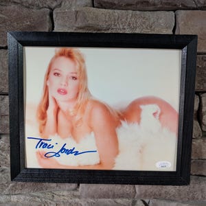 Puede incluir: Fotografía enmarcada que presenta a una persona con cabello rubio, vestida con una prenda blanca similar a piel. La imagen está firmada con tinta azul con el nombre "Traci Lords". La fotografía está encerrada en un marco negro.
