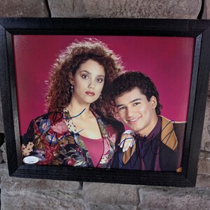 May include: Framed photograph featuring two people, a woman and a man, posing together. The woman wears a floral jacket over a pink top. The man wears a dark shirt with a gold and black design. Both have autographs.