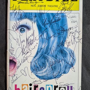 May include: A Playbill for the musical Hairspray, featuring a photo of a woman with blue hair. The Playbill is signed by several cast members, including Nikki Blonsky,  John Travolta, and  Harvey Fierstein. The Playbill is from the Neil Simon Theatre.