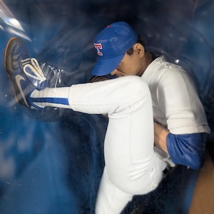 Op de afbeelding: Een honkbalfiguur van een pitcher in een blauw-wit uniform, met een blauwe pet met een rood "T" logo. Het beeldje staat in een werphouding, met één been uitgestrekt en het andere gebogen. De schoenen zijn blauw en wit.
