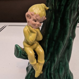 Pode incluir: Uma figura de cerâmica de um duende amarelo sentado em um tronco de árvore verde. O duende está vestindo uma roupa amarela e tem uma expressão triste no rosto.