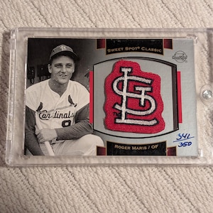 Può includere: Una carta da baseball con una foto in bianco e nero di Roger Maris, con una toppa con il logo dei St. Louis Cardinals rosso e bianco. La carta è racchiusa in un supporto protettivo trasparente. Il testo "SWEET SPOT CLASSIC" è visibile.