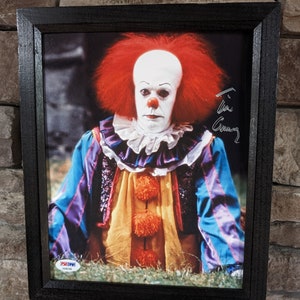 Op de afbeelding: Een ingelijste foto van een clown met rood haar en een wit gezicht. De clown draagt een kleurrijk kostuum met een geel, oranje en blauw patroon. De clown kijkt met een serieuze uitdrukking naar de camera. De foto is gesigneerd met "Tim Curry".
