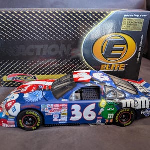 Op de afbeelding: Een blauw, rood en wit modelraceauto met nummer 36. De auto heeft verschillende sponsorlogo's en het M&M's-logo. Het is een 1:24 schaal Elite verzamelobject, met een doos op de achtergrond.
