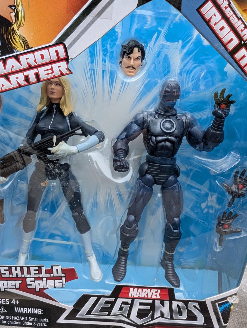 Puede incluir: Un set de figuras de acci&oacute;n Marvel Legends que incluye a Iron Man con un traje sigiloso y a Sharon Carter, una superesp&iacute;a de S.H.I.E.L.D. Las figuras est&aacute;n empaquetadas en un bl&iacute;ster de pl&aacute;stico transparente con fondo azul y blanco. El empaque incluye el logotipo de Marvel Legends y el texto "Stealth Iron Man" y "S.H.I.E.L.D. Super Spies!"