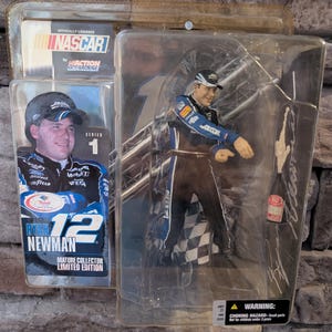 Op de afbeelding: Een NASCAR-actionfiguur van Ryan Newman in zijn #12 Alltel-auto. De figuur draagt een blauw-wit racepak met een zwarte helm. De figuur staat in een rennende houding met zijn rechterarm omhoog. De figuur is verpakt in een doorzichtige plastic blisterverpakking met een kartonnen achterkant. De achterkant toont een foto van Ryan Newman en de tekst "NASCAR by Action Mcfarlane Series 1 Ryan Newman #12 Mature Collector Limited Edition".