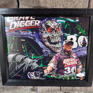 May include: Framed print featuring the Grave Digger monster truck. The image includes the Grave Digger logo, a skull design, and a man in a Grave Digger shirt. The print is framed in a black wooden frame.