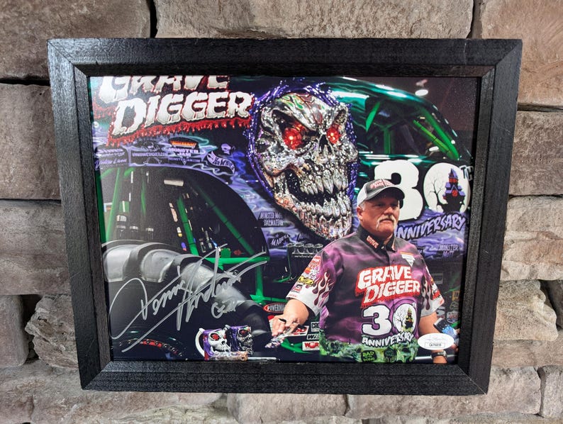 May include: Framed print featuring the Grave Digger monster truck. The image includes the Grave Digger logo, a skull design, and a man in a Grave Digger shirt. The print is framed in a black wooden frame.