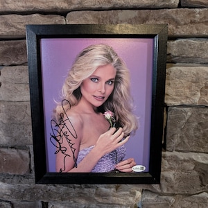 May include: Framed photograph of a woman with blonde hair, holding a pink rose, and wearing a lavender dress. The photo is signed and set against a purple background, enclosed in a black frame.