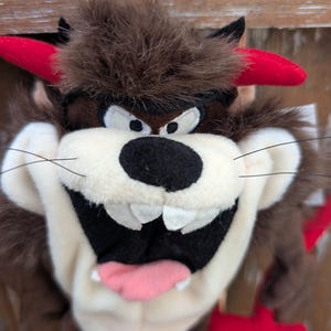 Könnte beinhalten: Ein Plüschtier des Tasmanian Devil, mit braunem Fell, roten Hörnern und einem breiten, zahnigen Grinsen. Der Teufel hat eine schwarze Nase, weiße Augen und eine rosa Zunge. Das Spielzeug hält einen roten Dreizack.