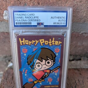 Puede incluir: Una tarjeta coleccionable de Harry Potter con una ilustración de Harry Potter en una escoba, con el texto "Harry Potter Quidditch Outfit". La tarjeta está en un estuche protector y está autografiada por Daniel Radcliffe.